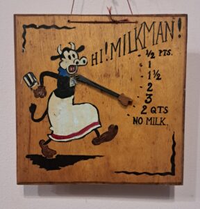 A wooden board with a painted image of a cow saying 'Hi Milkman!' and pointing to how much milk the family would like.