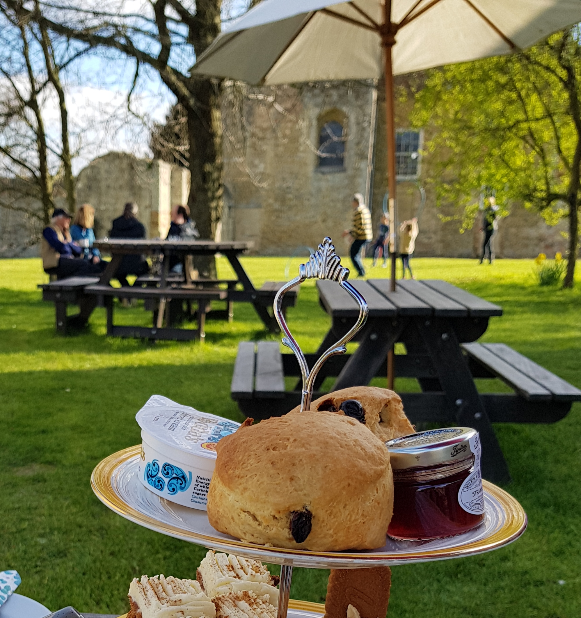 Scones, jam and cream on a plate at the front of the image. In the background, there are picnic benches, trees and a partial view of Denny Abbey.