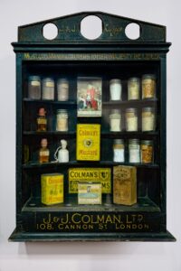 A black wooden cabinet with a glass front. Inside there are a number of yellow boxes and tins with the words 'Coleman's Mustard'