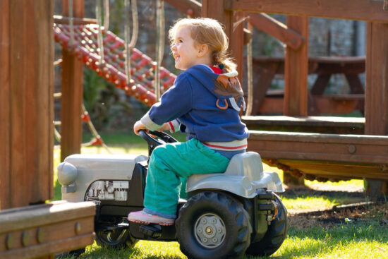 Girl riding a small, grey pedal tractor in a playground.