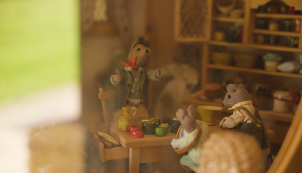 A close up image of a toy mouse wearing a waistcoat. It is surrounded by miniature models of food and living room furniture.