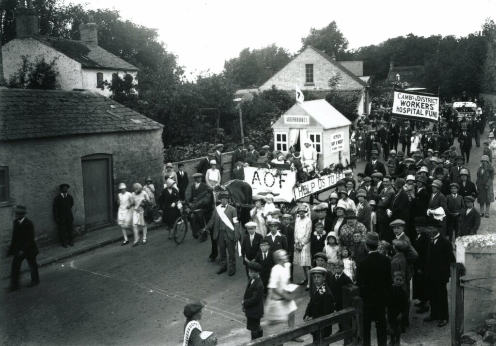 A black and white image, showing a parade through a village street