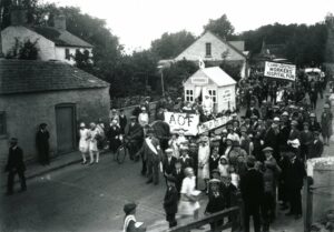 A black and white image, showing a parade through a village street