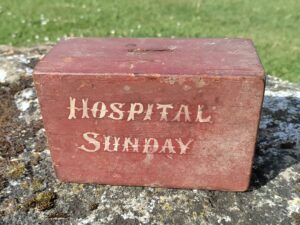 A small, red painted, wooden box with the words 'Hosptial Sunday' hand painted on the side in white