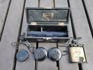 A black box that is open on a table with gold letters stating 'Mr R H Dent's Ardente for deaf ears'. In front of the box are two circular discs with speaker groves and another circular disc with five dots in the centre. On the right of these disks is a battery pack which is open revealing a flask shaped battery with the company name printed in the middle in red.
